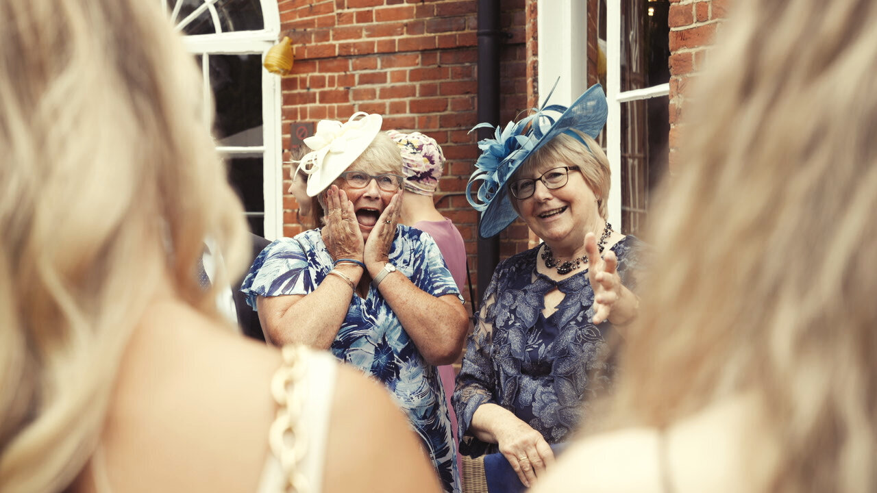 Two wedding guests in hats reacting with delight, one with her hands to her cheeks